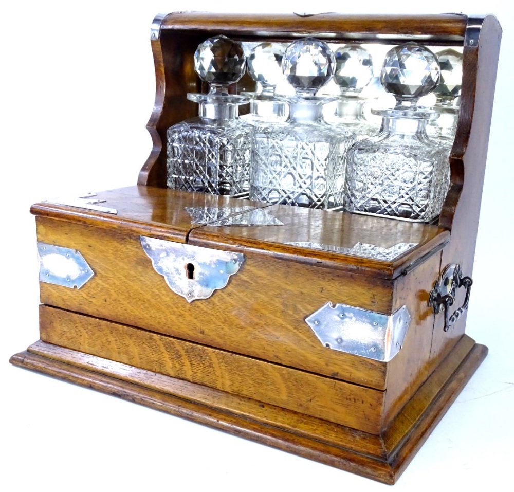 A late 19th/early 20thC oak and silver plated tantalus, with three square section decanters, a hinge