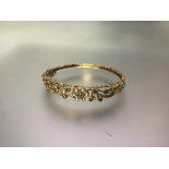 A 9ct gold hinged bangle set with emeralds and seed pearls, the leaf and scroll openwork mount