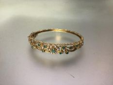 A 9ct gold hinged bangle set with emeralds and seed pearls, the leaf and scroll openwork mount