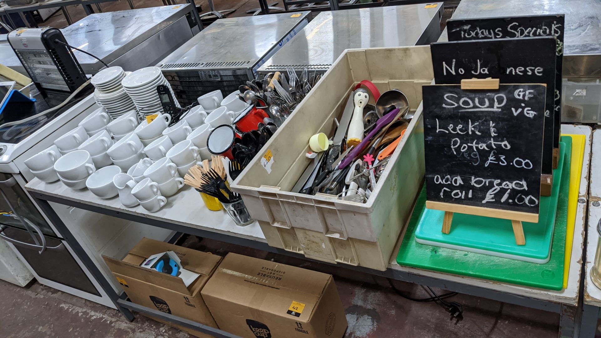 The contents of a table comprising white cups & saucers plus cutlery, utensils, chopping boards, min