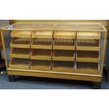 MID-CENTURY GLASS & LIGHTWOOD RETAILERS CABINET having a bank of sixteen tray-drawers, each with