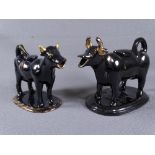 TWO BLACK JACKFIELD COW CREAMERS, one large and the smaller with gilt decoration, each on oval bases