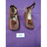 A pair of brown leather baby shoes having pom-pom's to the fronts and strap with button fastening.
