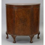 A reproduction burr walnut demi-lune side cabinet, fitted two shelves enclosed by a centre panel