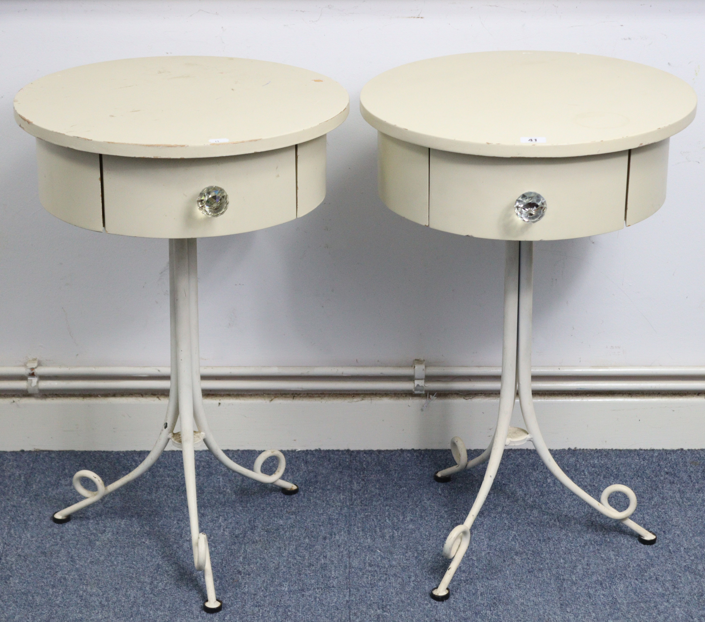 A pair of cream finish bedside tables, each with circular top, fitted frieze drawer, & on white