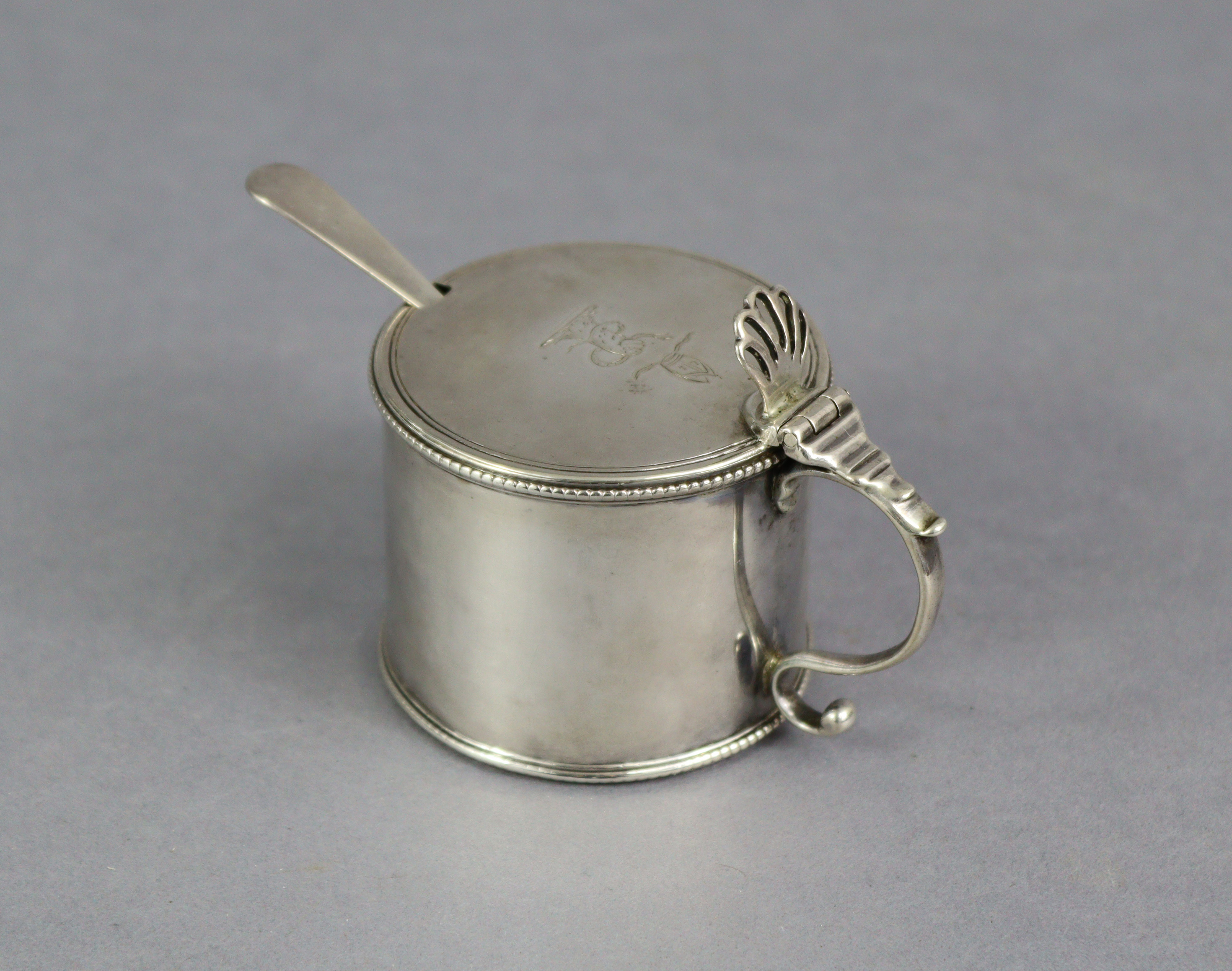 A George III silver drum-shaped mustard pot with pierced shell thumb-piece to the flat hinged lid,