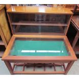 A teak bijouterie table with a hinged glazed top on square legs together with a teak bookcase with