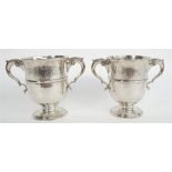 A pair of silver twin handled cups, each body decorated with a central band, with scrolling handles,