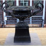 A black painted cast iron twin handle urn, on square stand, 63cm wide x 61cm high.