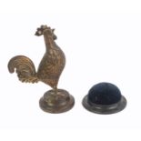 Two pin cushions, comprising a gilded brass example in the form of a standing cockerel with