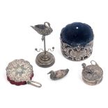 Three silver pin cushions, a ring stand and a tape measure, comprising a small silver duck form