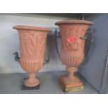 Two terracotta urns with cherub ornament on a pedestal foot, cast brass handles and a plinth