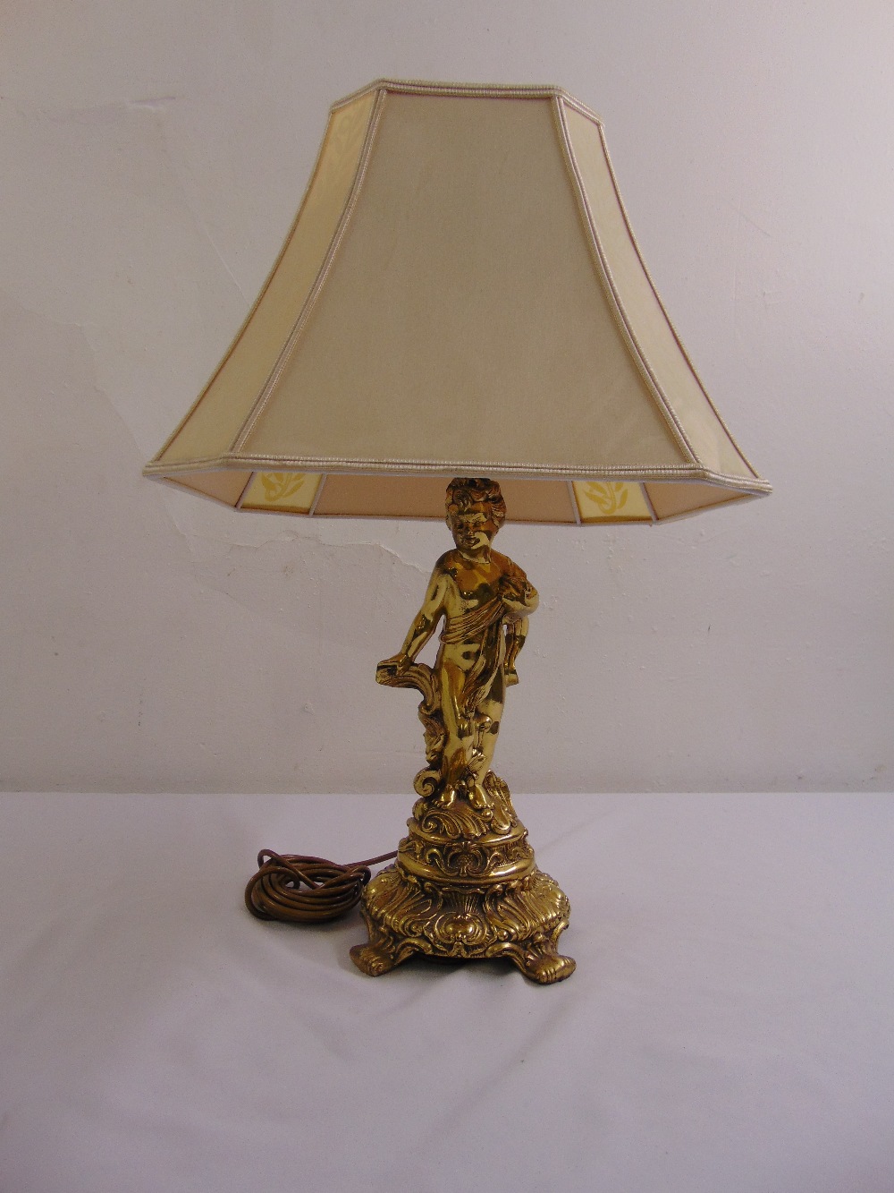 A gilded metal table lamp in the form of a putti on raised circular base with silk shade