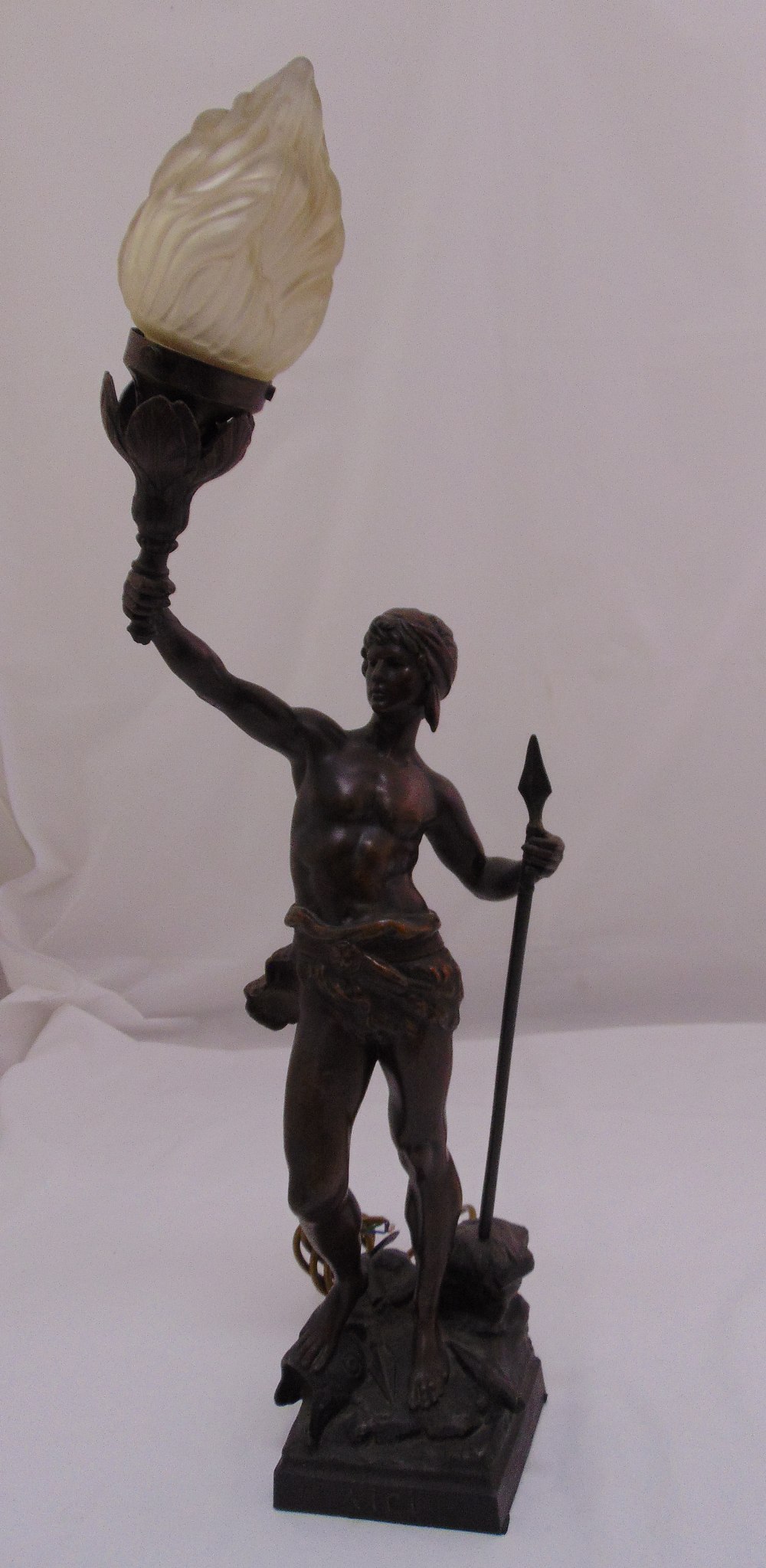 A spelter lamp stand in the form of a warrior holding a spear and supporting a frosted glass flame