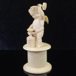 A 19th century carved ivory figure of Eros moulding a heart on an anvil, on drum shaped plinth base,