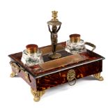 A William IV tortoiseshell inkstand, with central putto candlestick, pen tray to each side,