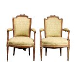 A pair of Louis XVI style upholstered fruitwood fauteuils, each crestrail headed by a trophy of