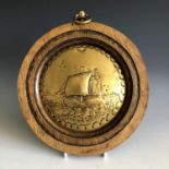 A small brass roundel in the manner of Obed Nicholls, decorated with a ship, mounted in an oak