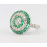 A platinum, emerald and diamond target dress ring set with a brilliant cut diamond surrounded by
