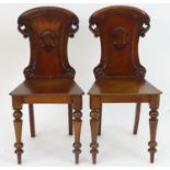 A pair of 19thC mahogany hall chairs with a carved backrest and armorial decoration above a