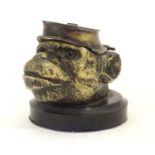 A Victorian novelty brass desk inkwell formed as the head of a monkey with glass eyes wearing a hat.