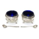 A pair of circular silver table salts, each raised on three hoof feet, with blue glass liners and