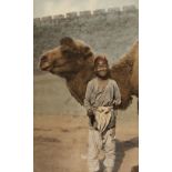 * China. A smiling boy holding the tether of his camel, hand-coloured carbon print, China, 1920s