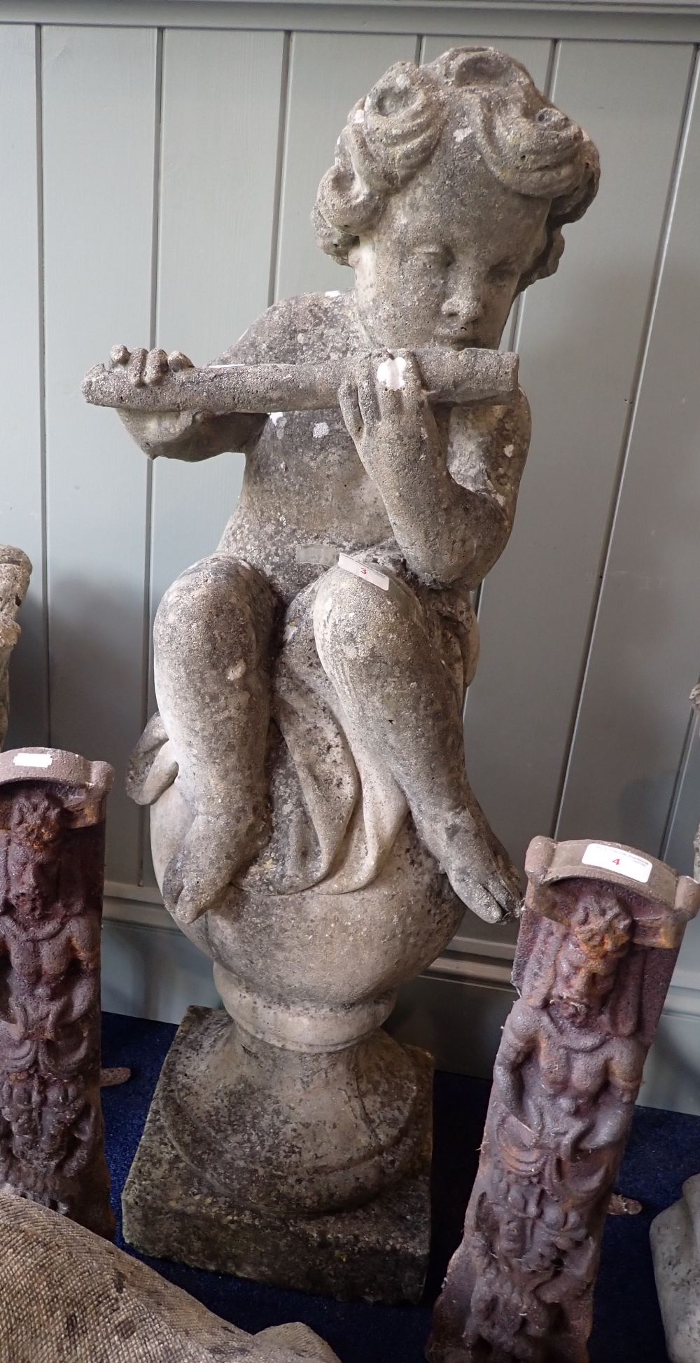 A RECONSTITUTED GARDEN ORNAMENT in the form of a Putto playing a flute sitting on a ball 100cms high