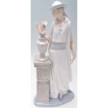 A Lladro porcelain figurine of a 1920's flapper girl modelled next to a jardiniere stand. Stamped