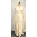A wedding dress, 1937, of ivory satin, bias-cut with cowl neck and ribbon-lace sleeve heads, a