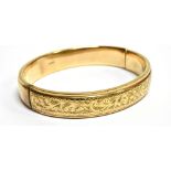 A 9 CARAT GOLD HINGED BANGLE scroll patterned front, plain reverse, of hollow construction, 9