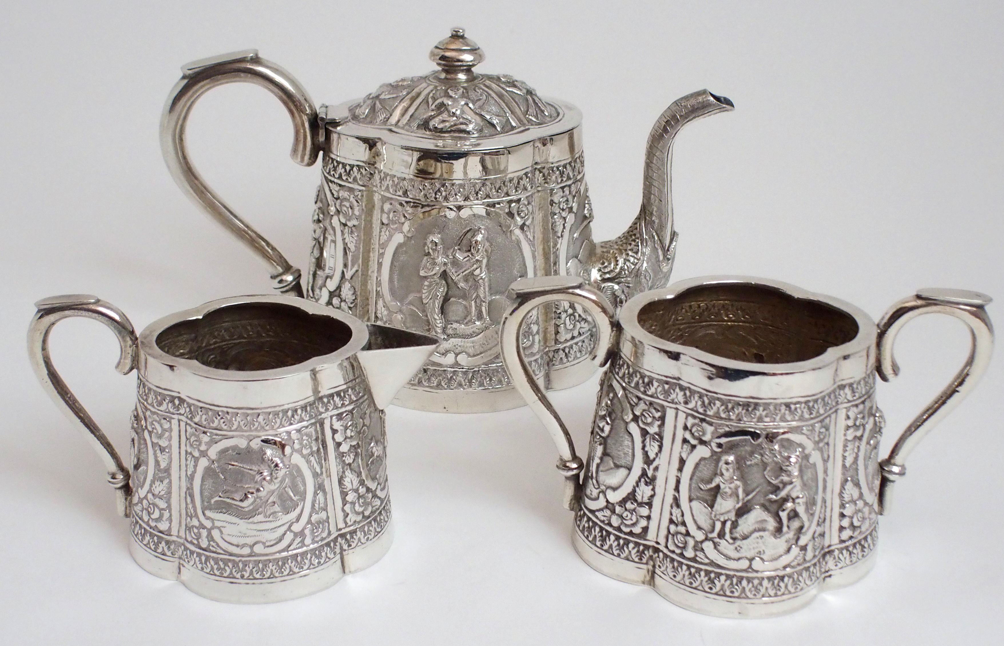 AN ASIAN WHITE METAL THREE PIECE TEA SERVICE cast and decorated with figures in cartouche panels