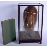 A TRIBAL SHRUNKEN HEAD together with a book within a glass case. Head 34 cm high.