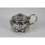 A George IV silver mustard circular form with lid and scroll handle, embossed floral decoration,