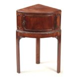 A GEORGE III MAHOGANY CORNER BEDSIDE TABLE with hinged door above a carved moulded edge, standing on