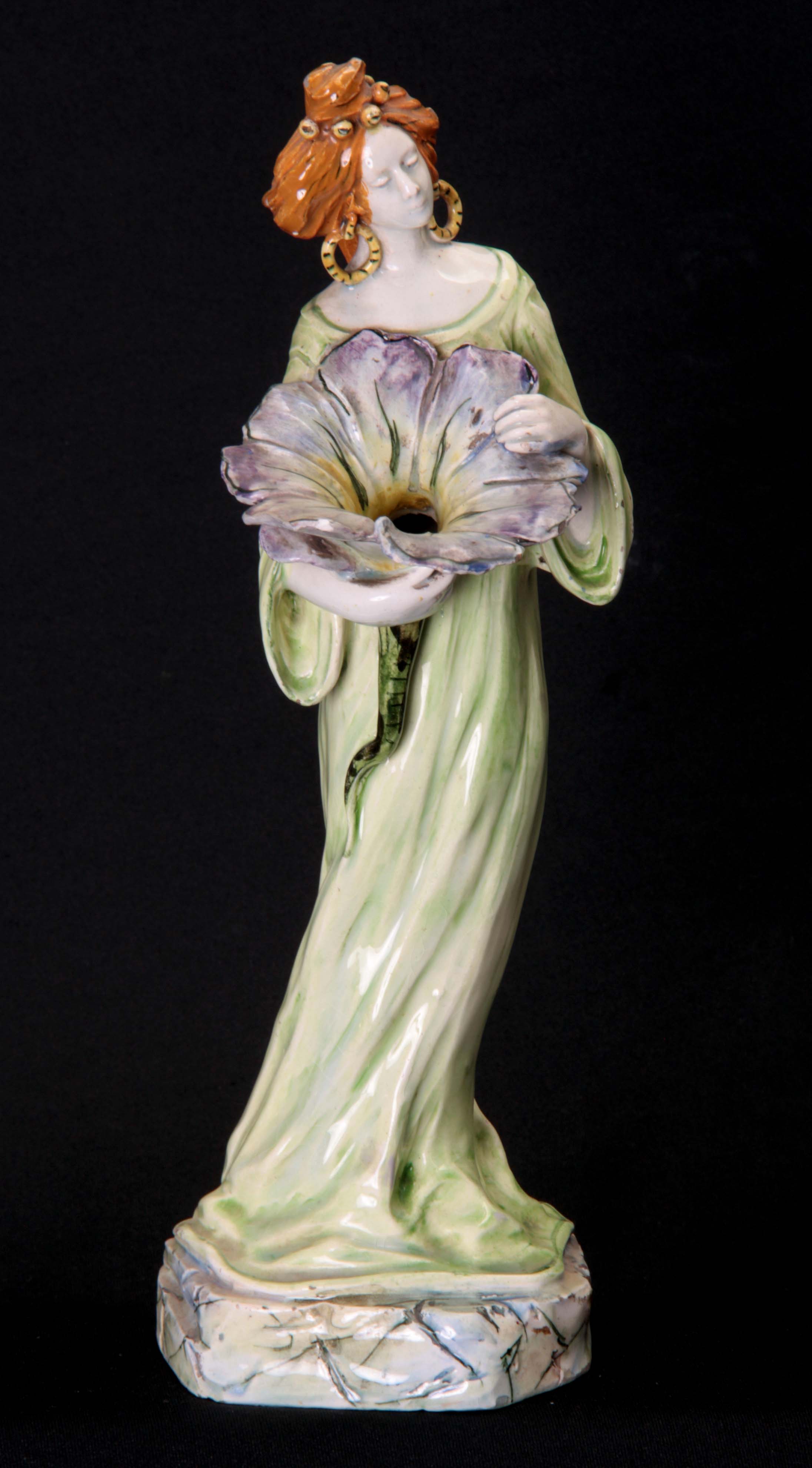 AN ART NOUVEAU GLAZED POTTERY FIGURE modelled as a standing lady clutching a flower head with hollow