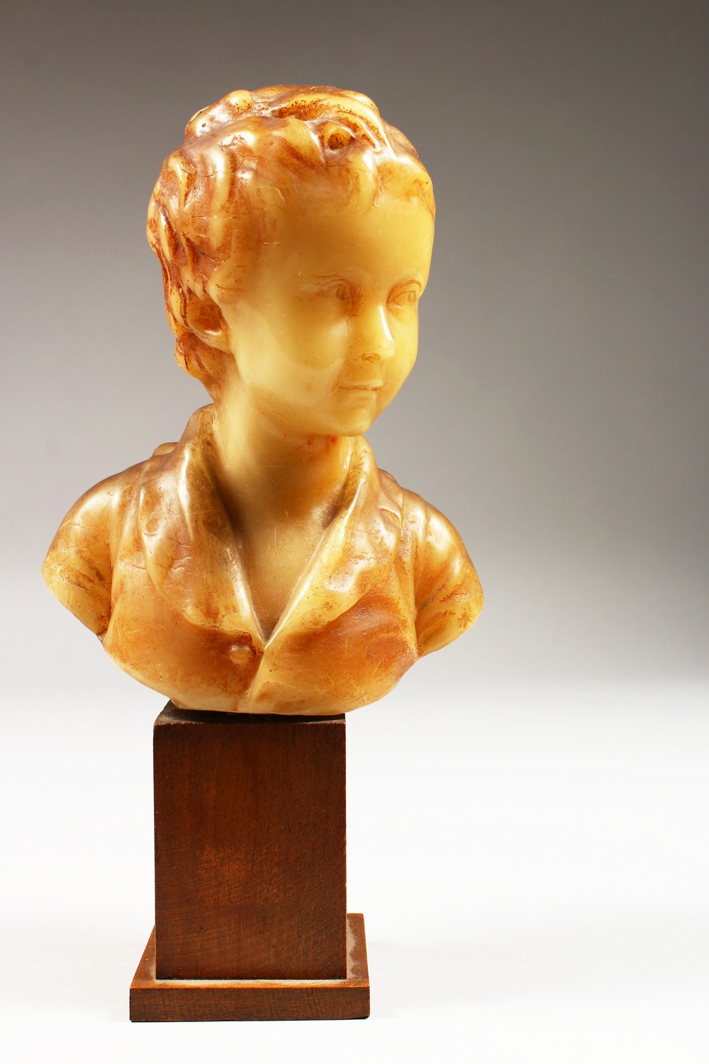 A SMALL GEORGIAN CARVED WAX BUST, head and shoulders of a young boy. 6ins high on a wooden plinth.