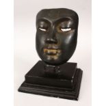 AN EASTERN BRONZE MASK with dark patination on a wooden base, 22cm high on stand, the mask 16cm high
