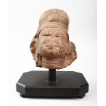 AN INDIAN RED SANDSTONE CARVING OF THE HEAD OF A DIETY, on a wooden stand, the stone 17cm high, on