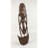 A CARVED WOOD TRIBAL "FISH HOOK", as a male figurine with shell eyes. 26.5ins high.
