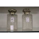 A pair of cut glass square shaped decanters with hinged embossed silver tops (one lacking stopper