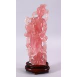 A CHINESE CARVED ROSE QUARTZ FIGURE OF GUANYIN, stood holding a bouquet of flora, on a carved wooden