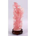 A CHINESE CARVED ROSE QUARTZ FIGURE OF GUANYIN, stood holding a bouquet of flora, on a carved wooden