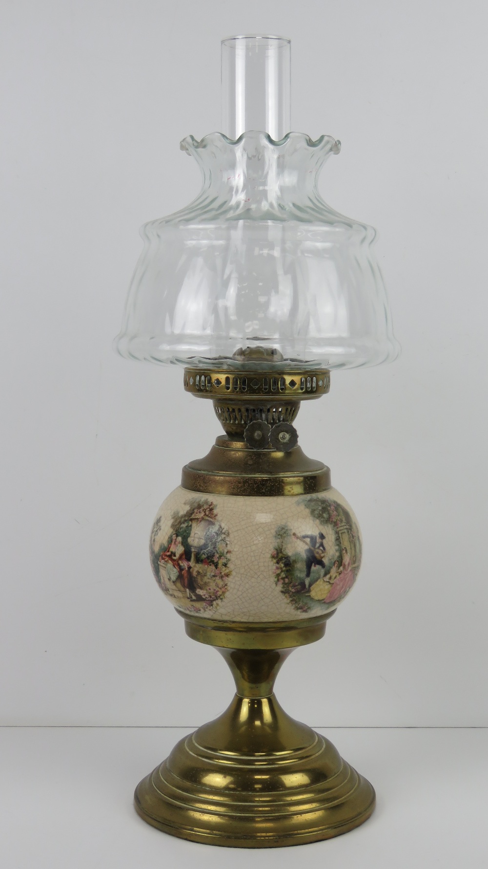 A brass oil lamp having ceramic reservoir with continental figural garden scenes upon,