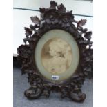 A VICTORIAN CARVED BLACK FOREST OVAL EASEL, BACK FRAME DECORATED WITH LEAVES AND FLOWERS. CONTAINING