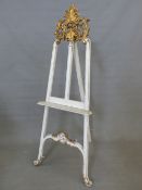 A WHITE PAINTED FLOOR STANDING EASEL WITH GILT FOLIAGE CRESTING AND SCROLL FEET, THE PICTURE REST. W