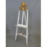 A WHITE PAINTED FLOOR STANDING EASEL WITH GILT FOLIAGE CRESTING AND SCROLL FEET, THE PICTURE REST. W