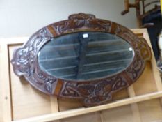 AN OVAL ARTS AND CRAFTS BEVELLED GLASS MIRROR IN A MAHOGANY FRAME CARVED WITH FOUR PANELS OF ENTRE