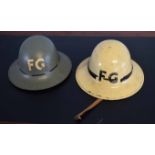Two vintage firemen's tin hats, each initialled FG, one grey and one beige