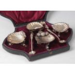 A late Victorian cased set of four silver shell shaped table salts, raised on dolphinium supports;